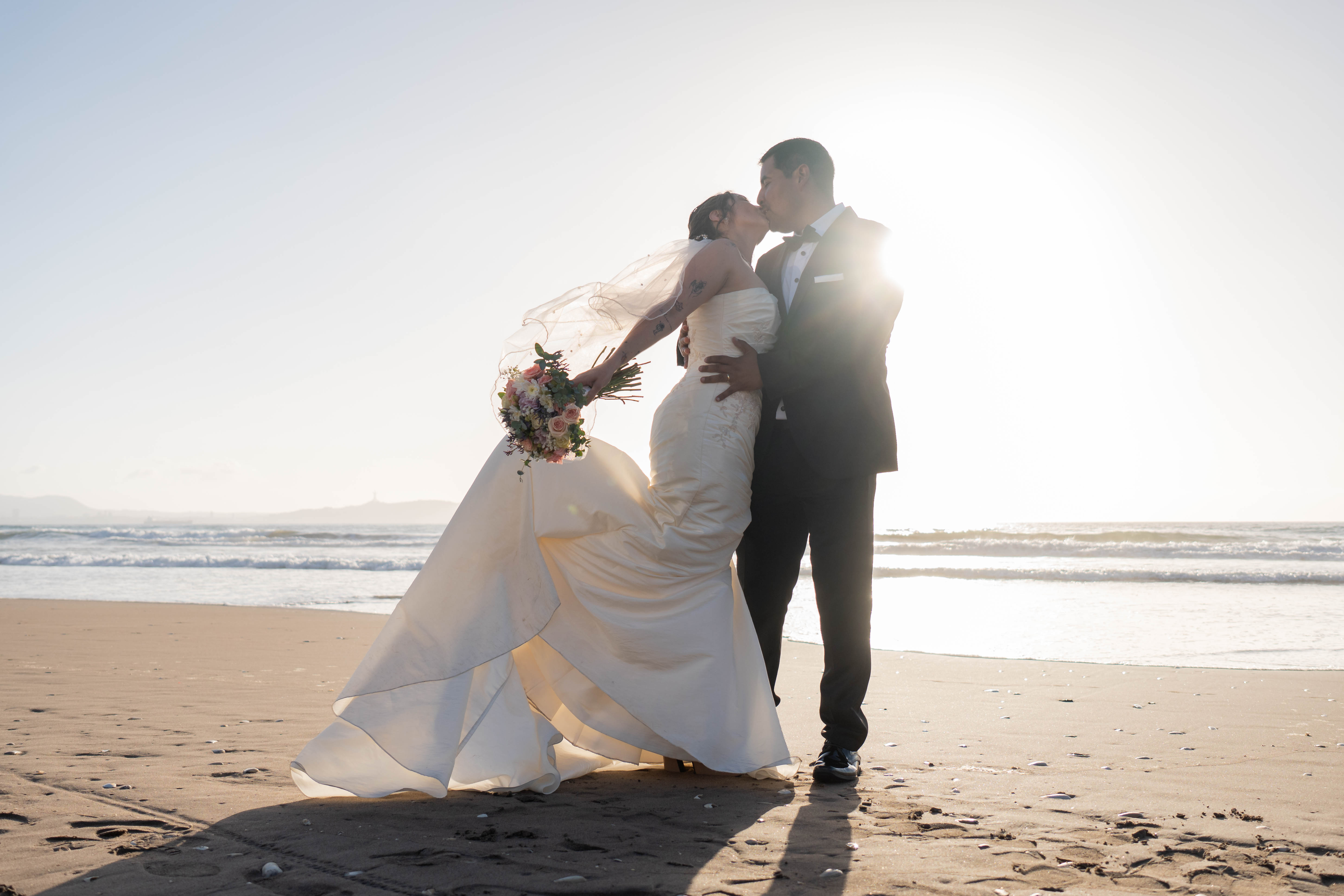 Un momento romántico capturado cuando los novios se besan en la playa al atardecer.