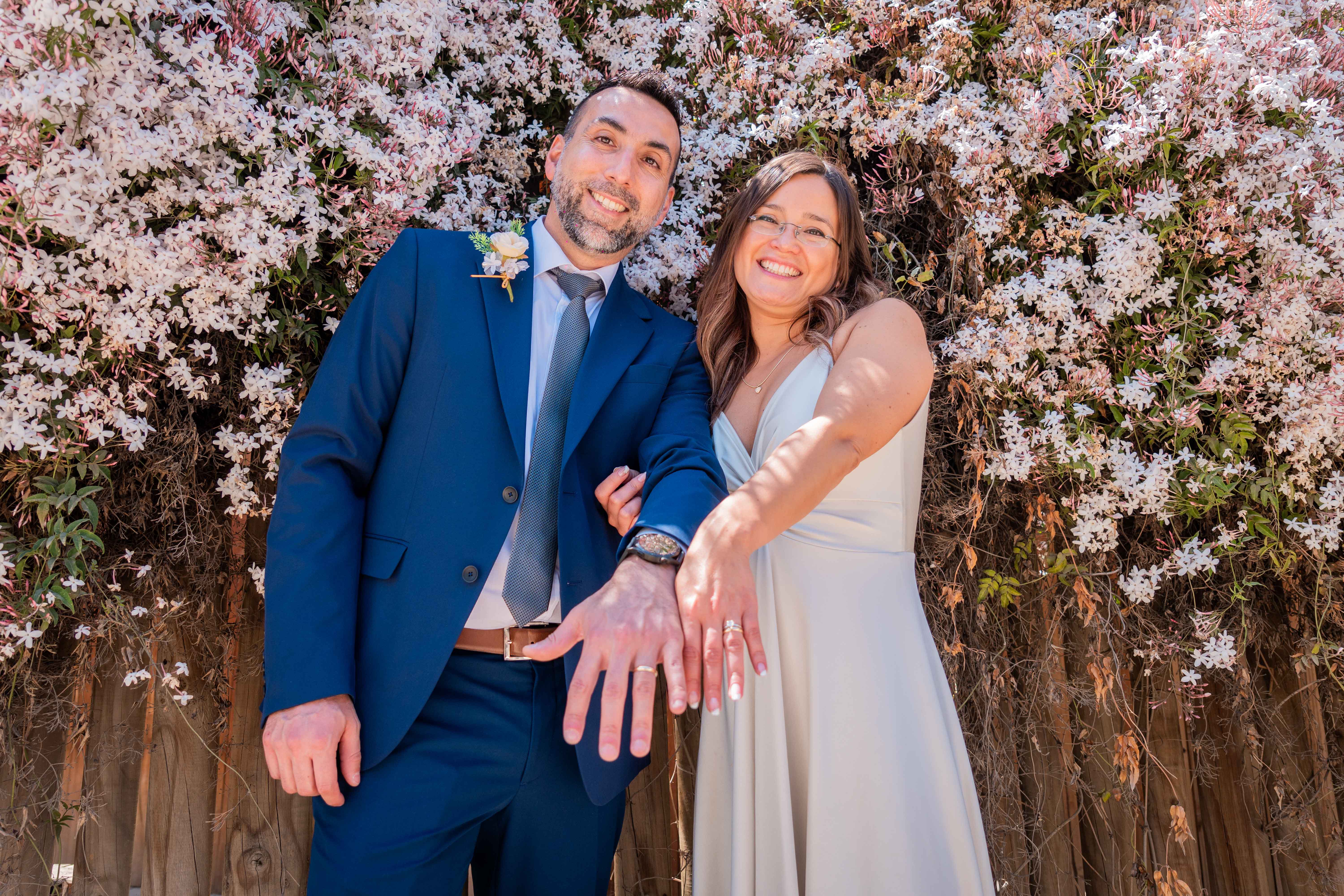 Una pareja de recién casados posando frente a un fondo flores rosadas y mostrando sus anillos de matrimonio en el día de su boda.
