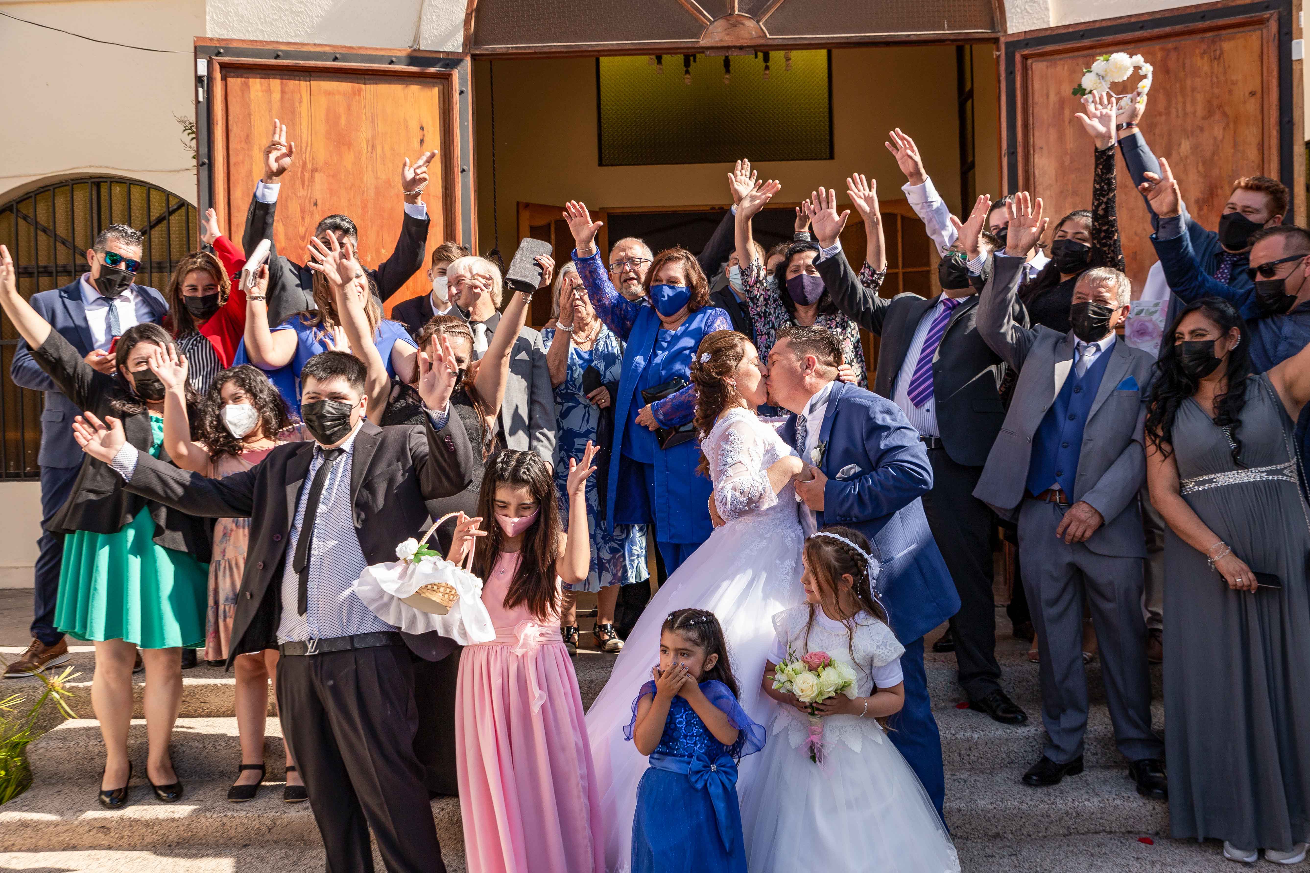 Un grupo invitados a la boda paradas frente a una iglesia, levantando la mano al unísono para celebrar a los novios.