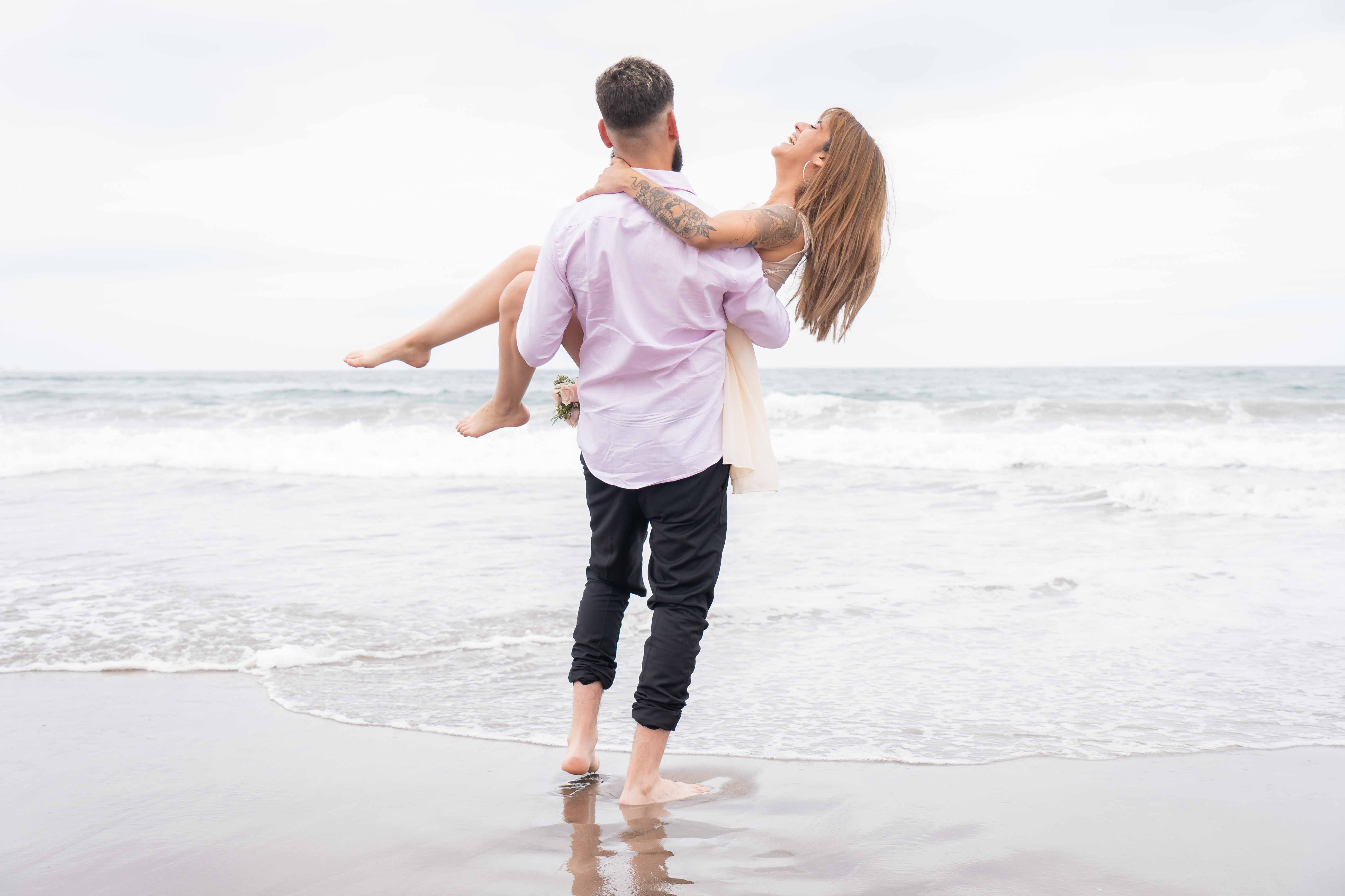 Hemoso momento de un novio cargando a su amada en por la orilla del mar en el día de su boda.