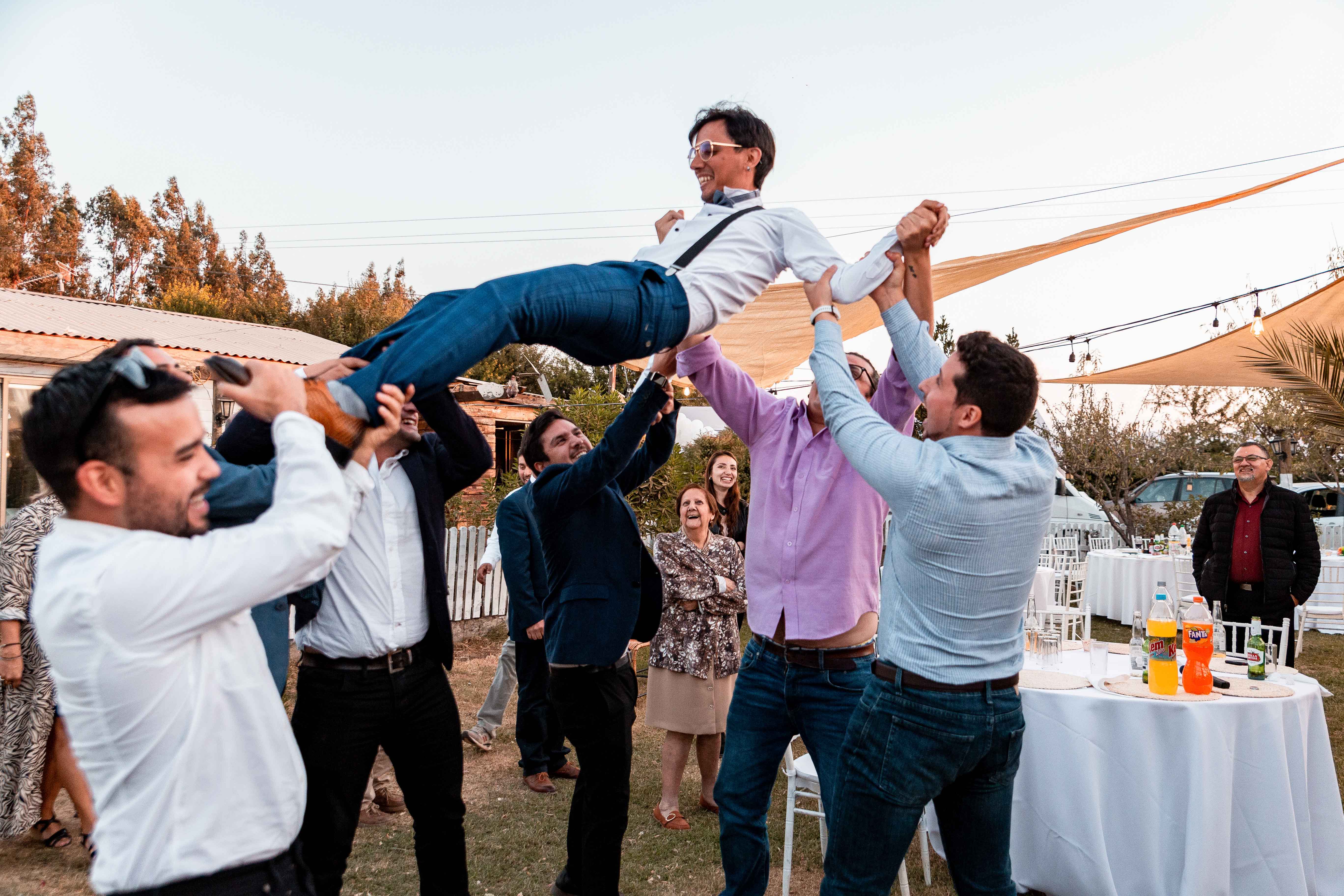 Grupo de amigos riendo, celebrando y cargando al novio en el día de su matrimonio.